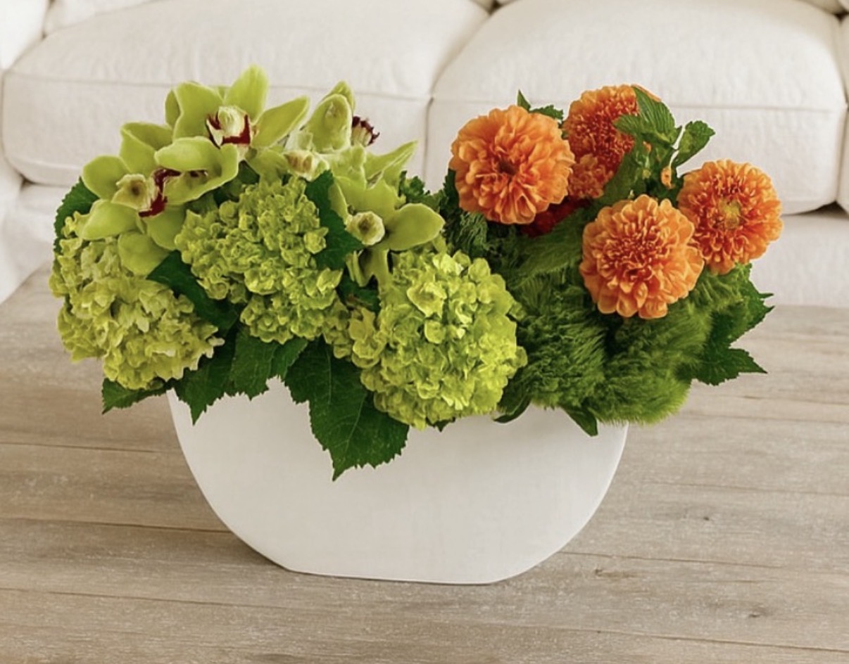 Bright orange dahlias, green hydrangeas and cymbidium orchids in a low white oval vessel