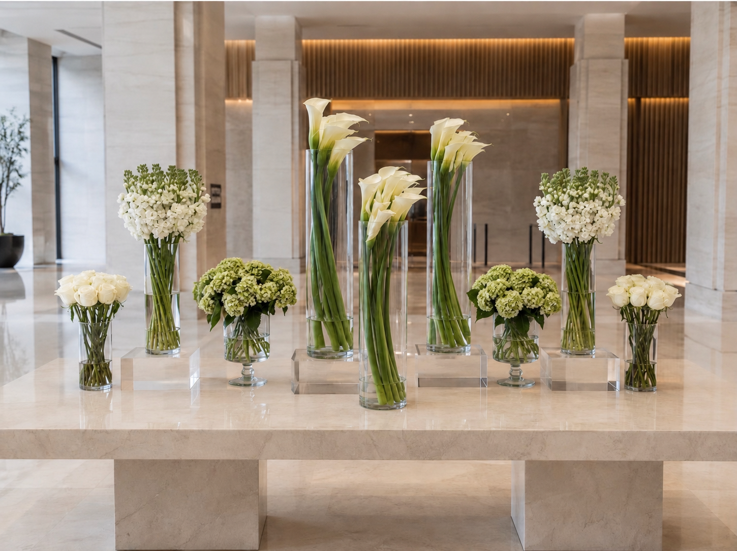 Symmetrical installation of white calla lilies, roses, and pale green hydrangeas in clear glass vessels on a stone lobby table